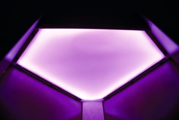 Eine geometrische Deckenleuchte strahlt ein sanftes violettes Licht aus und beleuchtet die schrägen Holzpaneele darunter. Die Leuchte hat eine sechseckige Form und erzeugt einen Farbverlauf von hellem Violett zu dunklen Rändern.