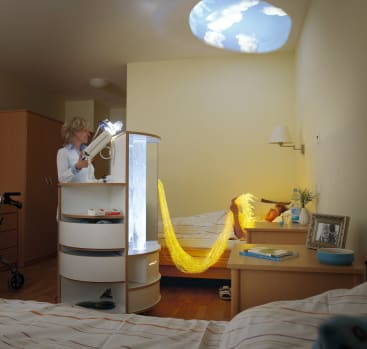 Eine Person steht neben einem Krankenhausbett, über das beleuchtete Glasfaserstränge drapiert sind. Ein Projektor zeigt ein Bild eines bewölkten Himmels an der Decke. In dem Raum sind eine Gehhilfe und medizinisches Material zu sehen.