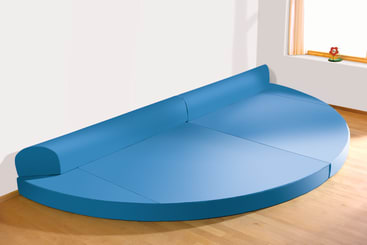 Ein halbrundes blaues Schaumstoffsofa mit Rückenlehne steht vor einer weißen Wand in einem Raum mit Holzfußboden. An der rechten Wand ist ein kleines Fenster mit einem Vorhang und einer roten Blumendekoration zu sehen.