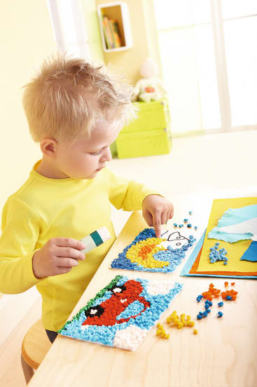 Ein kleines Kind mit blondem Haar sitzt an einem Tisch und gestaltet bunte Mosaikkunst mit kleinen Schaumstoffstücken. Das Kind trägt ein gelbes Hemd und konzentriert sich auf die Anordnung der Teile in einem blau-gelben Muster. Auf dem Tisch liegen Malutensilien.