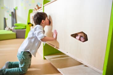 Ein kleines Kind kniet auf dem Boden und blickt durch ein rundes Loch in einer Holzplatte, während das Gesicht eines anderen Kindes durch eine sechseckige Öffnung in derselben Platte zu sehen ist. Der Schauplatz scheint ein Spielzimmer zu sein.