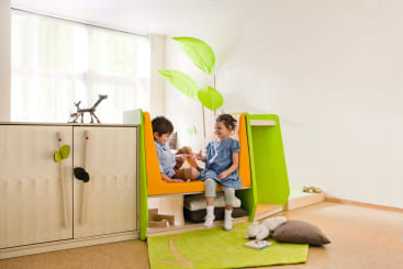 Zwei kleine Kinder sitzen und unterhalten sich in einer kleinen, farbenfrohen Spielecke mit grünen und gelben Tafeln. Der Raum ist mit Spielzeug, einem weichen Teppich, einem Schrank und großen Fenstern ausgestattet, die natürliches Licht hereinlassen.