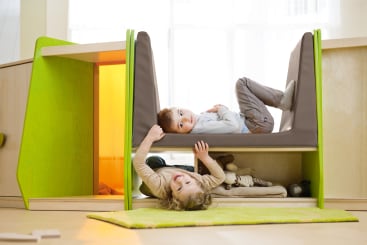 Zwei kleine Kinder spielen auf einem modernen Indoor-Spielgerät; eines liegt oben auf einer gepolsterten Plattform, während das andere darunter liegt und lächelt. Die Struktur ist grün und aus Holz, und darunter sind Stofftiere zu sehen.
