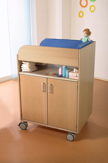 Ein hölzerner Wickeltisch mit einer blau gepolsterten Matte, einer Puppe, gefalteten Handtüchern, Babypflegeprodukten und Schränken darunter, die auf Rädern in einem hell erleuchteten Raum stehen.