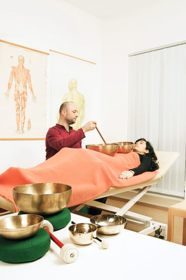 Eine Person, die auf einer mit einer roten Decke bedeckten Massageliege liegt, erhält von einem Therapeuten eine Klangtherapie-Sitzung mit Klangschalen. An der Wand im Hintergrund hängen anatomische Schautafeln.