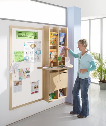 Eine Person steht an einer hölzernen Informationsstation mit Regalen mit Zeitschriften und Broschüren. An der Station befindet sich eine Pinnwand mit verschiedenen Aushängen und Flyern. Neben dem Fenster im Hintergrund ist eine Topfpflanze zu sehen.