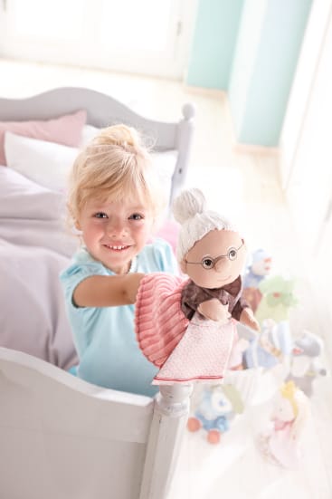 Ein kleines Kind mit blondem Haar steht lächelnd an einem weißen Bett und hält eine Puppe hoch, die wie eine ältere Frau mit Brille und grauem Haar aussieht. Im Hintergrund sind mehrere Plüschtiere und Spielzeuge zu sehen.