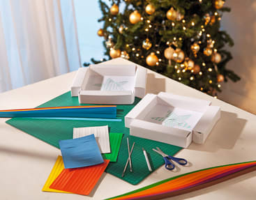 Ein Tisch mit bunten Quilling-Papierstreifen, zwei laufenden Quilling-Projekten auf weißen Tabletts, Pinzetten, Scheren und Bastelwerkzeug. Im Hintergrund steht ein geschmückter Weihnachtsbaum mit Ornamenten.