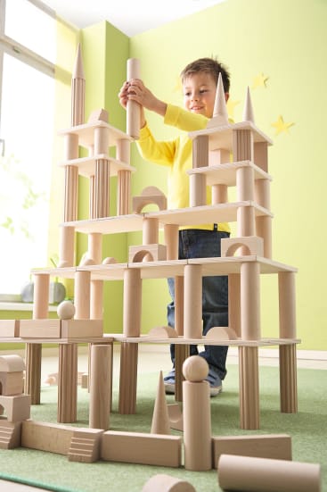 Ein kleines Kind in einem gelben Hemd steht drinnen und baut ein hohes, kompliziertes Bauwerk aus Holzklötzen auf einem grünen Teppich, während Licht durch ein nahes Fenster fällt.