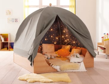 Ein hölzernes Indoor-Zeltbett mit grauem Baldachin, kuscheligen Kissen, Stofftieren, einer gelben Plüschsonne und sternförmigen Lichterketten im Inneren, das in einem hellen, organisierten Kinderzimmer mit Spielzeug und Bücherregalen steht.