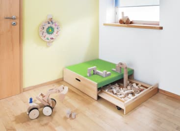 In einer hellen Ecke steht eine Holzspielzeugkiste mit einer grünen Spielfläche und einer offenen Schublade, die mit Holzklötzen gefüllt ist. Auf dem Boden und auf der Fensterbank stehen Holzfahrzeuge und Bauwerke. Ein verziertes Tamburin hängt an der Wand.