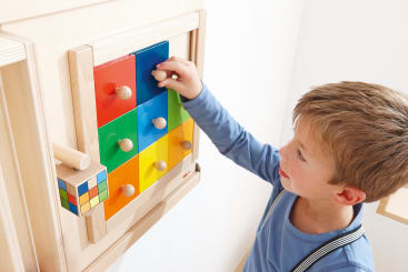 Ein Junge in blauem Hemd und Hosenträgern spielt mit einem an der Wand befestigten Holzpuzzle, das bunte Kacheln mit Knöpfen enthält. An der Seite der Holzstruktur ist ein kleiner Rubik's Cube angebracht.