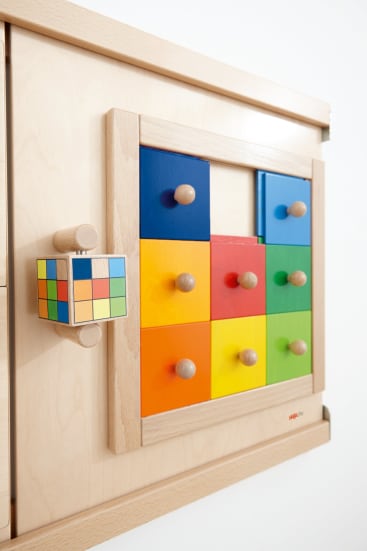 Hölzernes Wandpuzzle mit bunten quadratischen Fliesen und runden Knöpfen, die in einem Gitter angeordnet sind, neben einem kleinen Würfel mit bunten Quadraten. Der Hintergrund ist eine helle Holzoberfläche und eine weiße Wand.