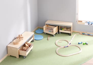 Ein aufgeräumtes Spielzimmer mit hellen Holzbänken, blauen Bodenkissen und Holzspielzeug, darunter eine Holzeisenbahn, die auf einem grünen Teppich in der Nähe eines Fensters mit natürlichem Licht aufgebaut ist.