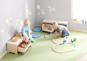 Zwei kleine Kinder spielen in einem hellen Raum mit grünem Teppich. Ein Kind sitzt in der Nähe eines mit Spielzeug gefüllten Holzwagens, während das andere eine Spielzeugeisenbahn auf einer auf dem Boden aufgebauten Holzschiene schiebt. In der Nähe befinden sich blaue Kissen und Spielzeuge.