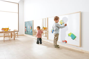 Zwei kleine Kinder spielen in einem hellen Klassenzimmer; eines malt an einer großen Tafel, während das andere in der Nähe herumläuft. Tische, Stühle und Spielzeug stehen im Hintergrund, und große Fenster lassen natürliches Licht herein.