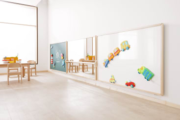 Ein helles Klassenzimmer mit hellem Holzboden, kleinen Holztischen und -stühlen und einer mit bunten Fahrzeugmagneten verzierten Tafel. Große Fenster lassen natürliches Licht herein.