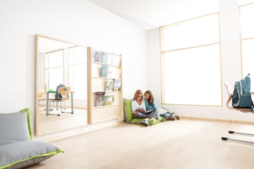 Zwei Kinder sitzen auf grünen Bodenkissen in einem hellen Raum mit großen Fenstern und lesen gemeinsam ein Buch. In der Nähe stehen Regale mit Büchern, ein Spiegel, ein Stuhl und Rucksäcke. Der Raum ist minimalistisch und modern gestaltet.