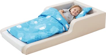 Ein kleines Kind liegt in einem beigen Kleinkindbett, das mit einer blauen Bettdecke und einem Kissen mit weißen Wolken und Schafsmotiven bedeckt ist. Das Kind lächelt und wirkt entspannt.