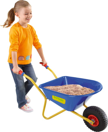 Ein junges Mädchen in einem orangefarbenen langärmeligen Hemd und Jeans schiebt eine blaue Spielzeugschubkarre, die mit Sand gefüllt ist. Die Schubkarre hat gelbe Griffe, ein schwarzes Rad und rote Griffe. Der Hintergrund ist weiß.