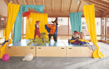 Drei Kinder in Kostümen treten auf einer kleinen, mit gelbem und blaugrünem Stoff dekorierten Bühne auf. Zwei Kinder sitzen auf der rechten Seite, während eines mit einem anderen in der Mitte der Bühne interagiert. Luftballons und Requisiten sind auf dem Boden verstreut.
