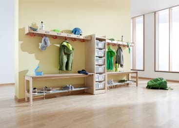 Ein heller Raum mit hölzernen Fächern, Haken und Bänken. Hier werden Jacken, Schuhe, Brotdosen und Helme der Kinder aufbewahrt. In durchsichtigen Plastikschubladen werden weitere Gegenstände aufbewahrt. Die Fenster auf der rechten Seite lassen natürliches Licht herein.