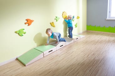 Zwei kleine Kinder spielen in einem hell erleuchteten Raum mit Holzboden. Eines klettert auf einem weichen Spielgerät, während das andere die Wanddekorationen in Form einer Sonne und von Vögeln berührt. Grünes Gras säumt die Wand in der Nähe eines Fensters.