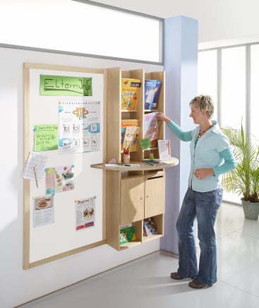 Eine Frau steht in der Nähe einer hölzernen Anschlagtafel und Informationsstation und blättert in Broschüren und Zeitschriften. Auf der Tafel sind Flugblätter, Aushänge und organisierte Literatur zu sehen. Im Hintergrund ist eine Topfpflanze zu sehen.