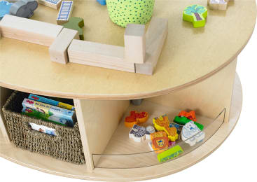 Ein hölzerner Spieltisch mit Bauklötzen und Tierfiguren obenauf, eine grüne gepunktete Tasse, ein geflochtener Korb mit Büchern und mehrere bunte Tierspielzeuge auf dem unteren Regal.