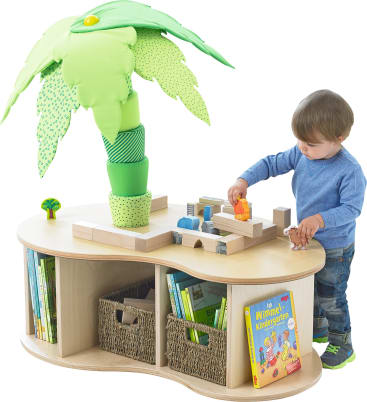 Ein kleines Kind spielt mit Holzspielzeug auf einem ovalen Tisch, in dessen Mitte eine große Plüschpalme steht. Unter dem Tisch befinden sich Regale mit Büchern, Körben und weiteren Spielsachen.