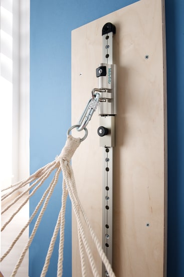 Nahaufnahme einer Hängemattenaufhängung, die auf einem vertikalen Holzbrett vor einer blauen Wand montiert ist, mit mehreren Seilen, die an einem Metallhaken und einer verstellbaren Schiene befestigt sind.