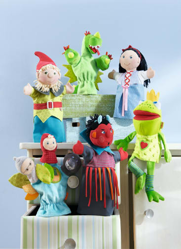 Sechs bunte Handpuppen, darunter ein Gnom, ein grüner Drache, ein Mädchen in einem blauen Kleid, ein Frosch, eine Hexe und eine Figur mit einem roten Umhang, sind auf Schubladen und Kisten vor einer hellblauen Wand angeordnet.
