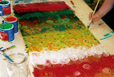 Eine Person malt bunte Strudel in Rot, Grün, Gelb und Weiß auf ein großes Blatt Papier, auf dem Tisch stehen Farbdosen, Pinsel und ein Glas Wasser.