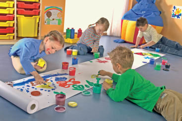 Vier kleine Kinder liegen auf dem Boden und malen gemeinsam mit bunten Farben und Pinseln auf einem langen weißen Blatt Papier. In einem hell erleuchteten Raum sind um sie herum Malutensilien und fertige Bilder zu sehen.