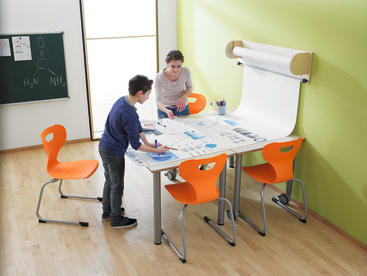 Eine Frau und ein Junge stehen an einem Tisch mit Zeichnungen und Papieren in einem Klassenzimmer. Um den Tisch herum stehen drei orangefarbene Stühle, und an der Wand ist eine Rolle weißes Papier angebracht. Im Hintergrund ist eine Kreidetafel mit Diagrammen zu sehen.