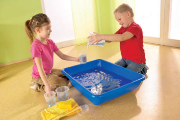 Zwei kleine Kinder sitzen in einem Haus auf dem Boden und schütten Wasser von einem Plastikbecher in einen anderen über einen großen blauen Plastikeimer. Mehrere Becher und gelbe Tücher stehen in der Nähe auf dem Boden.