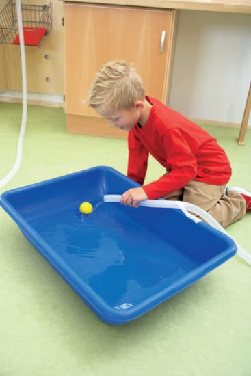 Ein Junge kniet neben einer großen blauen Plastikwanne, die mit Wasser gefüllt ist, und benutzt ein weißes Plastikwerkzeug, um einen gelben Ball zu führen, der auf der Wasseroberfläche schwimmt.