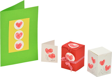 Eine grüne Grußkarte mit drei Herzformen, eine kleine weiße Karte mit zwei Herzen, eine rote Geschenkschachtel mit Band und Herzanhänger und eine weiße Schachtel mit Herzmotiven, alle vor einem weißen Hintergrund.