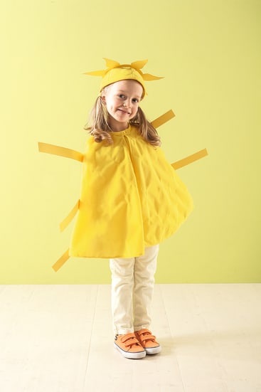 Ein kleines Kind steht vor einer hellgrünen Wand und trägt ein gelbes Kostüm, das an die Sonne erinnert, mit einem Stirnband in Form von Sonnenstrahlen, gelbem Klebeband für die Strahlen und orangefarbenen Turnschuhen. Das Kind lächelt und schaut in die Kamera.