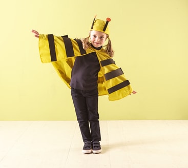 Ein Kind steht vor einer hellgrünen Wand und trägt einen gelben Umhang mit schwarzen Streifen und einen passenden Hut mit Fühlern, der an ein Bienenkostüm erinnert. Das Kind hat die Arme ausgebreitet und lächelt leicht.