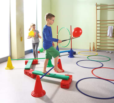 Zwei Kinder spielen in einem Turnraum mit bunten Kegeln, Reifen und Schwebebalken. Der Junge läuft barfuß auf einem Balken und balanciert einen roten Luftballon auf einem Stock, während das Mädchen mit einem gelben Ball hinterherläuft.