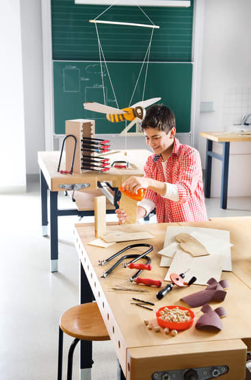 Ein Junge in einem rot karierten Hemd arbeitet mit einer Zwinge und einer Handsäge an einer Holzwerkbank in einem Klassenzimmer. Werkzeuge und Materialien liegen verstreut auf dem Tisch. Eine große Modellschere hängt von der Decke.