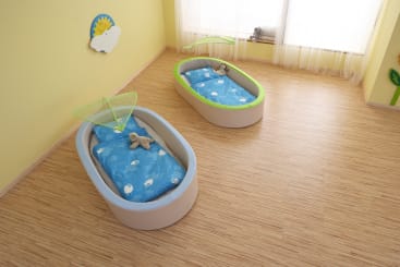 In einem hellen Raum mit gelben Wänden und einem Fenster, das von durchsichtigen Vorhängen verdeckt wird, stehen zwei ovale Kinderbetten mit blauer, wolkengemusterter Bettwäsche und Stofftieren auf einem Holzboden.