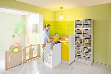 Ein Erwachsener hilft einem Kleinkind auf einem kleinen Wickeltisch mit Stufen in einem hell erleuchteten, grün-gelben Raum. In der Nähe befinden sich ein hoher Schrank mit Plastikschubladen und ein Laufstall mit einem Tor.