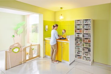 Ein Erwachsener steht an einer gelben Wickelkommode und wickelt ein Baby in einem hellen, modernen Kinderzimmer mit grünen Wänden, Schubladen mit Etiketten und einem durch ein Holztor abgetrennten Spielbereich.