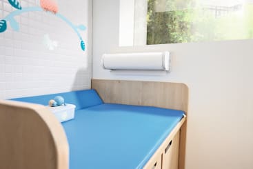Ein hölzerner Wickeltisch mit einer blau gepolsterten Matte steht neben einer Wand mit weißen Fliesen und einem dekorativen Mobile. Auf der Matte stehen eine Taschentuchbox und Spielzeug, und ein Fenster zeigt Grünzeug nach draußen.