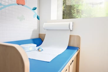 Eine Wickelkommode mit einer blauen Matte, einer herausgezogenen Rolle Einwegpapier, einem Behälter mit Feuchttüchern und einem kleinen Spielzeugball auf der Oberfläche, neben einem Fenster und einer gekachelten Wand mit einer Vogelgrafik.