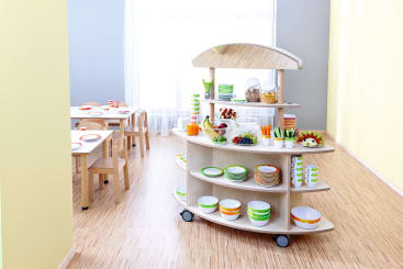 Ein hölzerner Essenswagen mit Regalen enthält Teller, Schüsseln, Tassen, Besteck, Müsli, Obst und Saft in einem hellen Raum. Im Hintergrund sind zwei kleine Tische mit Stühlen und Geschirr in der Nähe eines großen Fensters zu sehen.