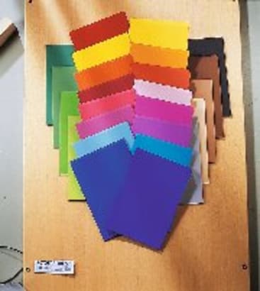 Eine Reihe von bunten rechteckigen Mustern, die fächerförmig auf einer hölzernen Oberfläche angeordnet sind und einen Farbverlauf von Gelb, Rot und Orange zu Grün, Blau, Lila, Rosa, Braun, Grau und Schwarz zeigen.