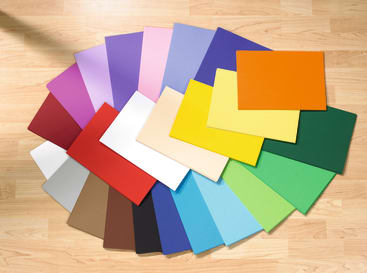 Verschiedene farbige Papierbögen sind in einem Halbkreis auf einem Holzboden angeordnet. Die Blätter umfassen eine breite Palette von Farben wie rot, blau, grün, gelb, orange, lila, braun, weiß und schwarz.